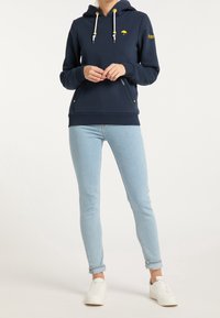 Felpa blu navy con tasca a marsupio, lacci gialli e un piccolo logo a forma di ombrello. Abbinata a jeans skinny azzurri chiari e scarpe da ginnastica bianche.