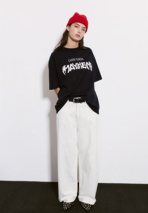 Fekete, oversized póló fehér grafikus szöveggel, széles fehér nadrággal és fekete, tűsarkú, szegecselt cipővel párosítva. Piros kötött sapka.