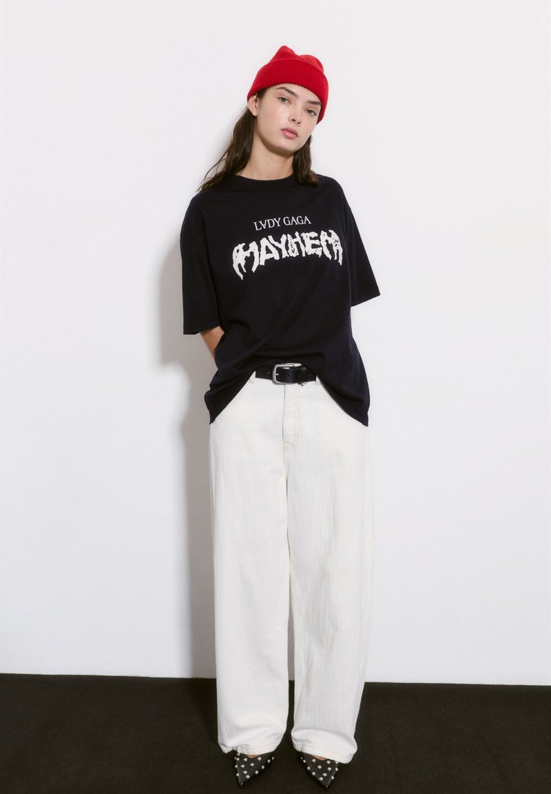 Mustat ylisuurikokoiset t-paidat, joissa on valkoista graafista tekstiä, yhdistettynä leveisiin valkoisiin housuihin ja mustiin niiteillä varustettuihin korkokengiin. Punainen neulottu beanie.