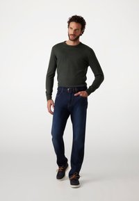 Dunkelgrünes Langarmshirt kombiniert mit blauen Jeans. Ausgestattet mit einem gewebten marineblauen Gürtel und lässigen marineblauen Schuhen mit braunen Schnürsenkeln.