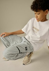 Jasnoszare spodnie cargo z czarnymi akcentami i naszywką z logo, w zestawie z białą koszulką z złotym napisem. Sneakersy w neutralnych kolorach.