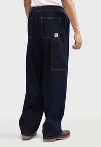 Dunkelblaue Jeans mit einem lockeren Schnitt, zwei großen Gesäßtaschen, gelben Nähten und einem elastischen Taillenbund.