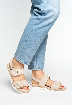 Pieds portant des sandales beige à boucle avec semelles blanches et vernis à ongles rouge, assortis à un jean bleu clair raccourci.