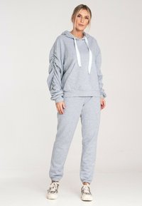Szara bluza z kapturem z białymi sznurkami, z akcentem na marszczone rękawy i pasującymi spodniami dresowymi, wykonanymi z miękkiego, teksturowanego materiału.