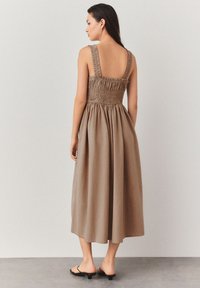 Braunes Midikleid mit einem gekräuselten Oberteil, breiten Trägern, offenem Rücken und fließendem Rock. Kombiniert mit schwarzen Sandalen. Glatter Stoff.