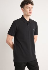 Zwarte polo met een zachte stof, voorzien van een twee-knopsluiting en een klein groen logo op de linkerkant van de borst. Eenvoudige, korte mouwen.