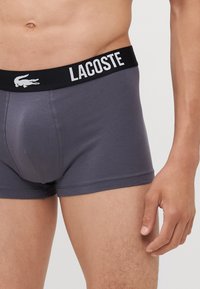 Sötét szürke boxer alsó, puha anyagból készült, széles fekete derékpánttal, amelyen a fehér "LACOSTE" logó és krokodil embléma található.
