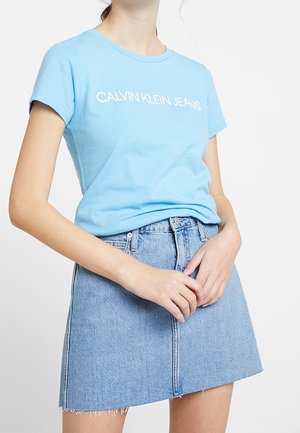 Jasnoniebieska bawełniana koszulka z nadrukiem "CALVIN KLEIN JEANS" w kolorze białym, zestawiona z wysokim stanem spódnicą dżinsową z postrzępionym brzegiem.