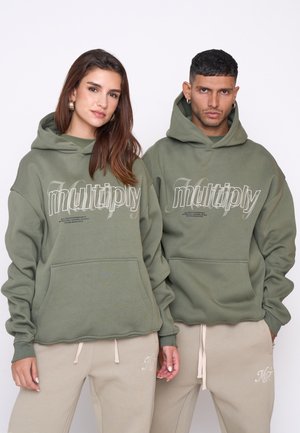 Felpe verde oliva con tasca frontale e design testuale. Abbinato a pantaloni tuta beige con ricamo. Vestibilità ampia, materiale morbido.