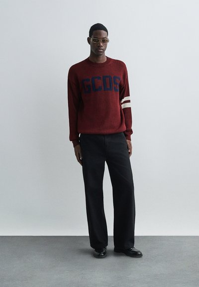 Rød strikkegenser med blå "GCDS"-logo, hvite striper på ermene, kombinert med løse svarte bukser og svarte sko, på en nøytral bakgrunn.