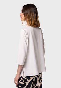 Blusa bianca leggera con maniche a tre quarti, scollo rotondo e spacchi laterali, abbinata a una gonna a fantasia nera e beige.