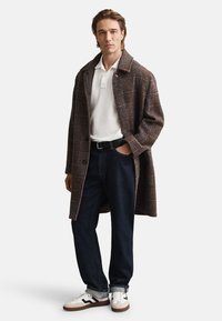 Brauner karierten Mantel über einem weißen Poloshirt, dunkelblaue Jeans und weiße Sneaker mit braunen Akzenten; weiche Textur, lockere Passform, lässiger Stil.