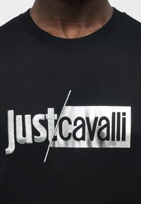 Άνδρας που φοράει μαύρο μπλουζάκι με στρογγυλή λαιμόκοψη και λευκό-μαύρο κέντημα με το λογότυπο "Just/cavalli" στο στήθος.