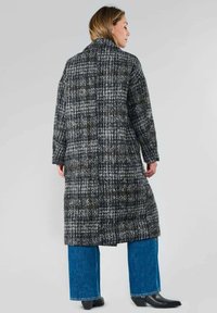 Manteau long à carreaux gris et noirs avec un col haut, en tissu texturé et avec une coupe décontractée, associé à un jean large bleu et des bottes noires.