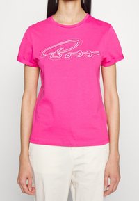 fuchsiové bavlněné tričko s rolovanými rukávy a bílým vyšívaným logem na přední straně, s klasickým kulatým výstřihem.