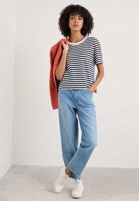 T-shirt rayé bleu marine et blanc avec un col rond, associé à un jean large bleu clair et des baskets blanches. Un cardigan orange tombe sur un bras.