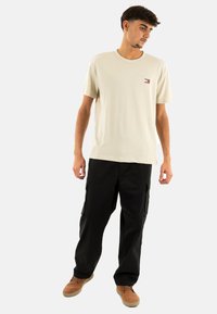 T-shirt in cotone beige con un piccolo logo sul petto, abbinato a pantaloni cargo neri e scarpe marroni. Si concentra su un fit casual e confortevole.