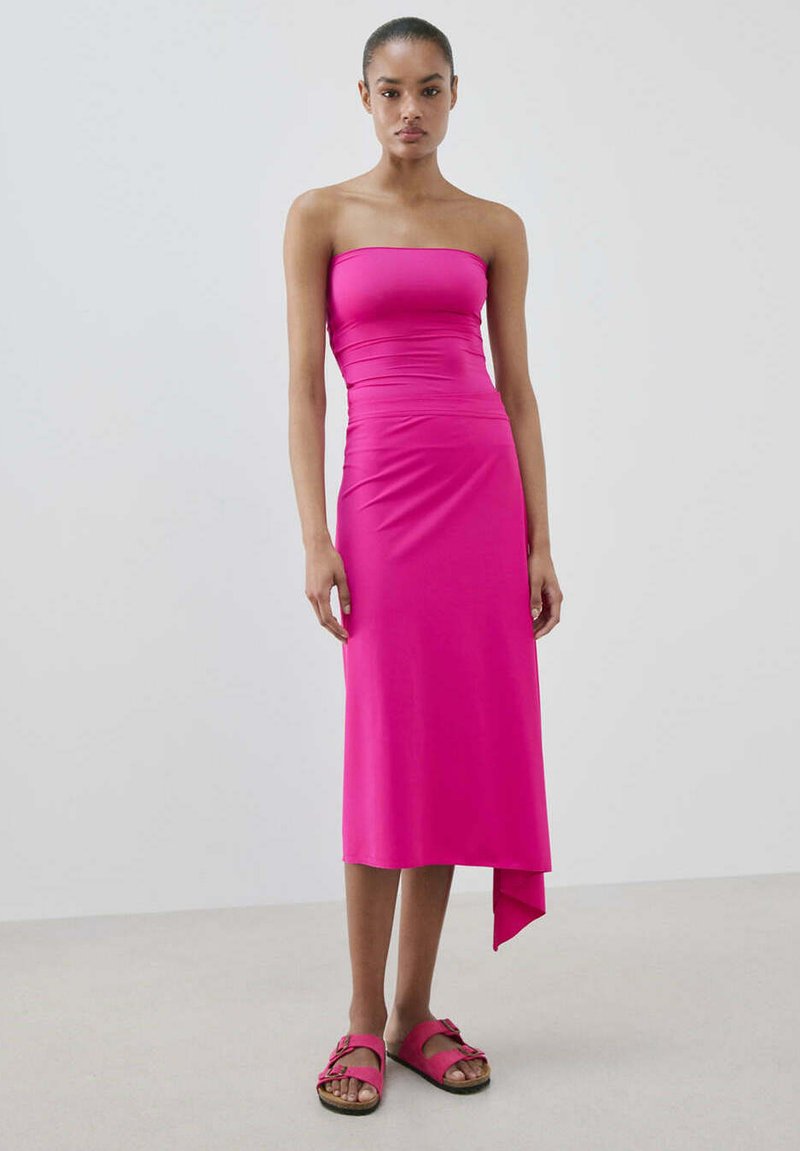 Femme debout portant une robe midi rose vif sans bretelles avec un ourlet asymétrique et des sandales roses assorties sur un fond uni.
