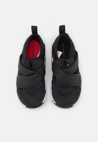 Black athletic shoes with a soft fabric upper, cross-straps in black rubber, and a red insole, featuring a white sole and no laces.