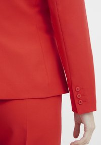 Veste de costume rouge en tissu lisse, coupe ajustée, détails aux poignets avec trois boutons. Design moderne et épuré.