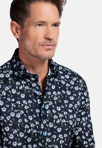Chemise florale bleue avec un fond sombre, présentant des marguerites blanches et des tiges vertes. Elle a un col classique et une fermeture à boutons.