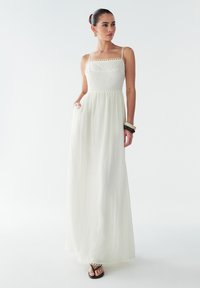 Maxi-jurk van doorschijnende witte stof met een aansluitend bovenstuk. Voorzien van spaghettibandjes, kant afwerking en een vloeiende rok. Gecombineerd met zwarte sandalen.