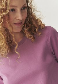 Purple cotton t-shirt with a round neckline, featuring a smooth texture and short sleeves, paired with loose, curly hair.