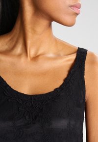 Krk, rameno a dolní část obličeje ženy, která na sobě má černou krajkovou tílko s propracovanou výšivkou na ramínkách a výstřihu.