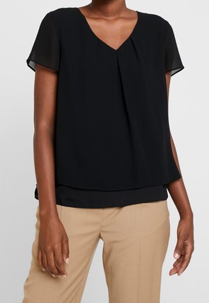 Frau trägt eine schwarze Bluse mit kurzen Ärmeln und V-Ausschnitt mit Falten-Detail, kombiniert mit beigen, maßgeschneiderten Hosen vor schlichtem Hintergrund.