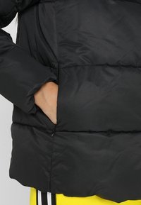 Chaqueta acolchada negra con un bolsillo con cremallera, diseño acolchado y una textura suave y ligeramente brillante. Detalles visibles de costura.