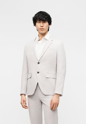 Mężczyzna w jasnoszarej marynarce i dopasowanych spodniach na białej koszuli, stojący na jednolitym jasnym tle.