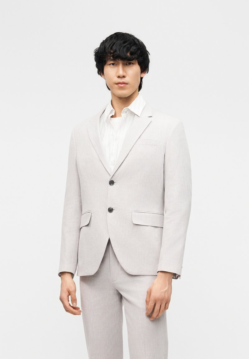 Άνδρας που φοράει ανοιχτογκρί σακάκι και αντίστοιχο παντελόνι πάνω από λευκό πουκάμισο, στέκεται μπροστά σε απλό φωτεινό φόντο.