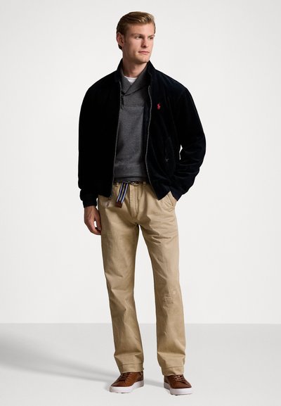 Giacca bomber in corduroy nera, maglione grigio, pantaloni beige, sneakers marroni con suole bianche e una cintura a strisce multicolori. Design pulito.