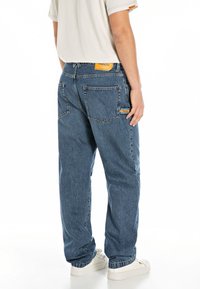 Relaxed-fit blue denim jeans with a mid-rise waist, standard pockets, and an orange brand patch on the back. Worn with white sneakers.