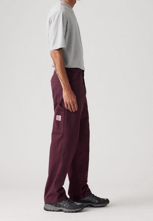 Homme portant une chemise grise à manches courtes, un pantalon cargo violet foncé avec une poche latérale, et des chaussures de sport noires, debout de profil devant un fond uni.