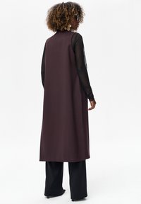 Długa, bezrękawnik w ciemnym fioletowym kolorze o prostym kroju, noszony na czarnej bluzce z długim rękawem i spódnicy z szerokimi nogawkami. Gładki materiał i minimalistyczny design.