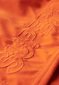 Tela naranja con una textura suave, destaca la letra en relieve "SUPER". La costura es precisa, añadiendo detalle al diseño.