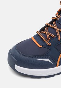 Sportschuhe aus dunkelblauem Stoff mit orangen Akzenten. Mit strukturiertem Gummisohle, gepolsterter Kragen und geflochtenen Schnürsenkeln. Reflektierende Details vorhanden.