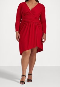 Červené zavinovací šaty s dlouhými rukávy a výstřihem do V. Design se zpevněným pasem, asymetrický lem, vyrobené z měkké, elastické látky. Černé boty na kotníkovém pásku.