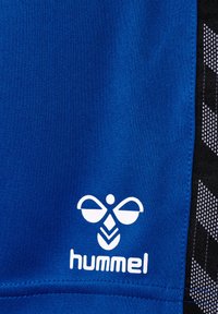 Blåt sportsstof med hvidt logo med abstrakte former og ordet "hummel." Sort tekstureret sidepanel med geometrisk mønster.