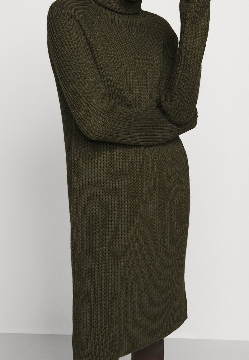 Femme portant une longue robe-pull en maille côtelée vert foncé à col roulé, posant avec un bras plié et la main près du visage.