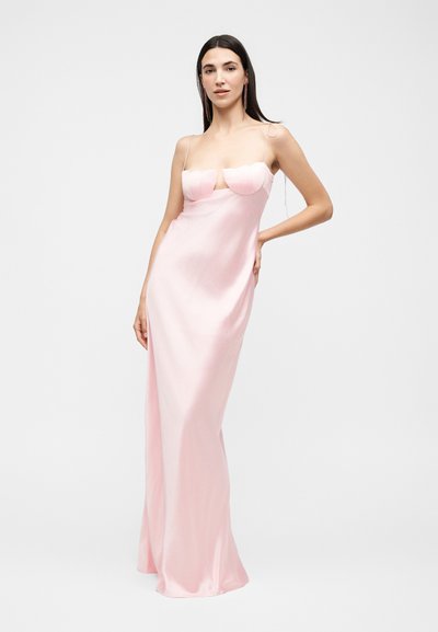 Γυναίκα στέκεται με ανοιχτό ροζ σατέν φόρεμα με λεπτές τιράντες και δομημένο μπούστο, ποζάροντας μπροστά σε απλό φωτεινό φόντο.