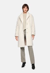 Weißer, plüschiger, übergroßer Mantel mit weitem Kragen, drei Knöpfen und seitlichen Taschen, kombiniert mit einem beigen Pullover und grauen Karohosen.