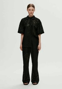 Μαύρο δυαίο σύνολο που περιλαμβάνει ένα χαλαρό, κοντομάνικο πουκάμισο και φαρδιά παντελόνια, και τα δύο με laser-cut λουλουδάτα σχέδια και κουμπιά μπροστά.