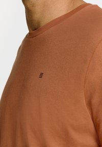 Bruine katoenen t-shirt met een ronde hals en korte mouwen. Heeft een subtiel zwart merkontwerp op de bovenste linkerkant. Zachte textuur.