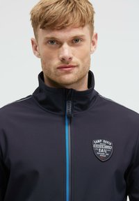 Chaqueta azul marino con cremallera y acabado texturizado, con una franja azul en la parte delantera y un logo de insignia en el lado izquierdo del pecho. Diseño de alto cuello.