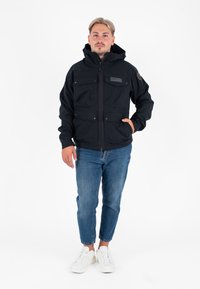 Man met licht haar in een zwarte rits-hoodie, blauwe spijkerbroek en witte sneakers, staand tegen een witte achtergrond.