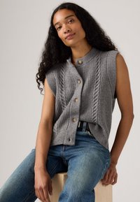 Grå ermeløs cardigan med ribbestruktur og kabelstrikkedesign, med rund krage og knappelukking, sammen med blå jeans.