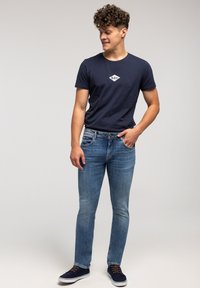 Młody mężczyzna z kręconymi włosami, ubrany w granatową koszulkę, jasnoniebieskie dżinsy i granatowe sneakersy, stojący i patrzący w bok.