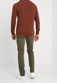 Mężczyzna stojący plecami, ubrany w sweter w kolorze rdzy, oliwkowe spodnie i białe sneakersy na jednolitym jasnym tle.